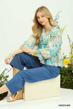 A woman wears an umgee watercolor floral blouse with sheer flounce sleeves in teal and green over blue pants