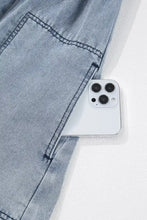 A white iphone 13 pro peeking from the front pocket of a vintage wash denim dress with pockets