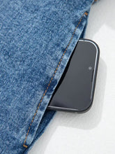 Black smartphone with silver trim peeking from front pocket of blue denim jeans worn with sleeveless denim dress