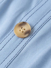 A round translucent gold-toned button with two holes on light blue woven fabric of a wide strap sweetheart neck denim dress